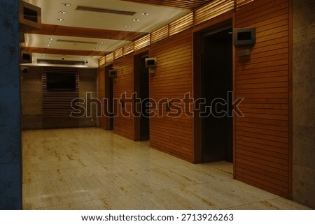 A modern elevator lobby featuring warm wood panel walls, soft ceiling lighting, and polished stone flooring. The minimalist design and symmetrical layout create a calm,                              
