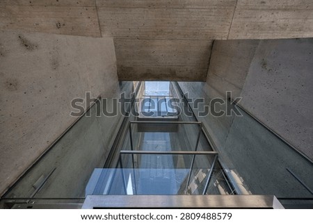 A modern elevator shaft featuring transparent glass walls framed with metal and surrounded by textured concrete surfaces in a contemporary architectural setting.