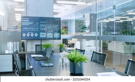 Modern Empty Meeting Room With Big Conference Table With Various Documents And Laptops On It, On The Wall Big TV Showing Company's Growth, Statistics And Pie Charts.