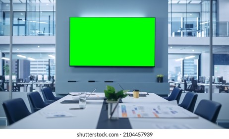 Modern Empty Meeting Room with Big Conference Table with Various Documents and Laptops on it, on the Wall Big TV with Green Chroma Key Screen. Contemporary Minimalistic Designed Workplace.