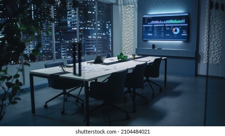 Modern Empty Meeting Room with Big Conference Table with Various Documents and Laptop on it. Wall TV Showing Company's Growth, Statistics, Graphs and Pie Charts. Late Evening or Night City Outside. - Powered by Shutterstock