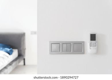 Modern Equipment And Save Energy Concept. Selective Focus On Minimalistic Light Switch And Air Conditioner Remote Controller On Copy Space White Wall Against Bedroom Interior At Blurred Background