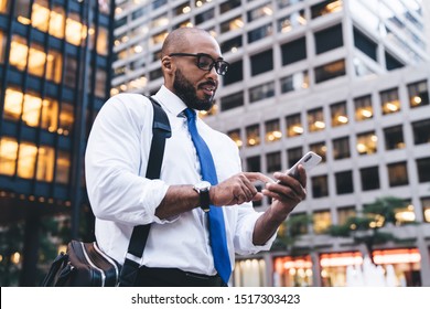Modern f African American bald bearded employee in glasses messaging on white smartphone while standing in front of modern office buildings