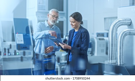 Modern Factory: Female Engineer, Male Project Manager Standing In High Tech Development Facility, Talking And Using Tablet Computer. Contemporary Facility With CNC Machinery, Robot Arm Production Line