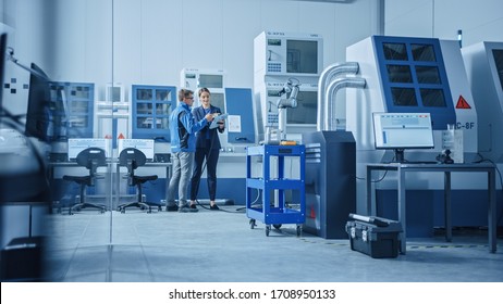 Modern Factory: Female Project Manager And Male Engineer Standing, Talking And Using Tablet Computer For Programming Robot Arm Assembly Line. Contemporary Facility With High Tech CNC Machinery