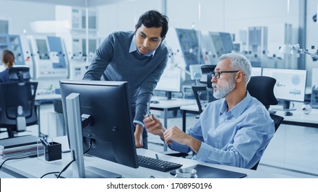 Modern Factory Office: Indian Project Manager Talks To Caucasian Senior Engineer Whos Working On Computer, Inspect Metal Component. Professionals Solving Heavy Industry Problems, Having Discussion