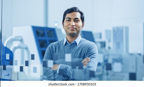 Modern Factory Office: Portrait Of Handsome Indian Engineer Crosses Arms And Smiles Charmingly. In The Background High Tech Facility With CNC Machinery, Robot Arm And People Working