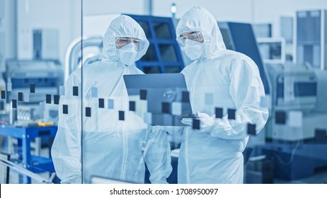 Modern Factory Sterile Workshop: Professionals In Coveralls, Masks Use Laptop And Talk. Medical Manufacturing Laboratory With High Tech CNC Machinery, Robot Arm Production Line And Automatic Equipment