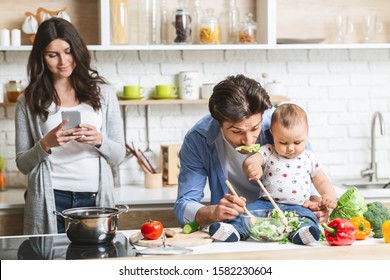 Modern family morning. Daddy cooking salad with baby son, mother networking on cellphone, kitchen interior - Powered by Shutterstock