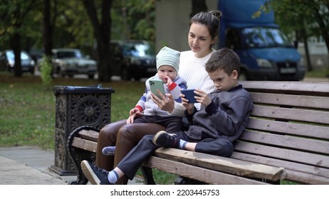 Modern Family Relaxing With Gadgets Outdoors On A Bench Outside. Mom And Two Sons In The Park.