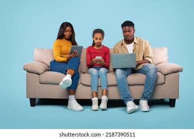 Modern Family. Young African American Father, Mother And Daughter Using Different Gadgets, Sitting On Couch Over Blue Studio Background