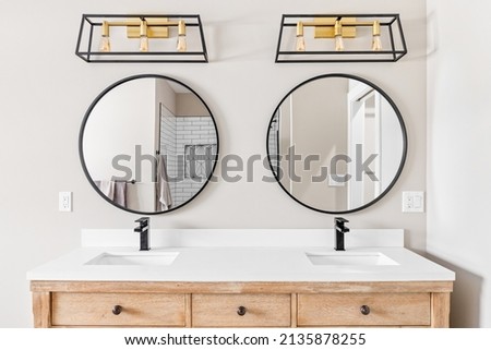A modern farmhouse bathroom with a natural wood vanity cabinet, black and gold light fixtures above circular mirrors, and granite countertop.