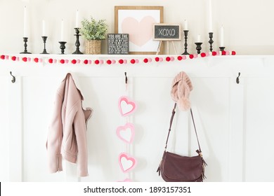 A modern farmhouse mantle decorated for Valentines Day. There is pink, red and white felt ball garland, a spring plant, black candle holders, a pink heart photo and clothes hanging from black hooks - Powered by Shutterstock