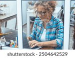 Modern female entrepreneur working from her home office on a laptop, seen through the window, representing the rise of small online businesses and the integration of technology in entrepreneurship