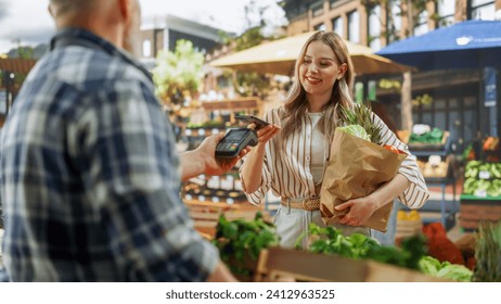 Modern Female Shopper Using Smartphone with Contactless Payment Technology to Pay for Organic Vegetables at a Farmers Market. Street Vendor Holding an Electronic Online Payment Terminal Device - Powered by Shutterstock