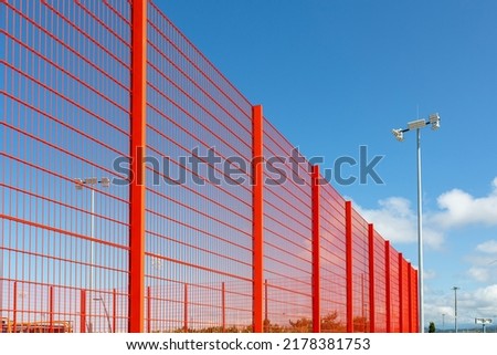 Modern fencing of the sports ground with a metal red fence. Lighting devices in the background