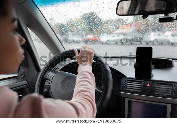 Modern gadgets for driving, working in city and\
traveling. Serious millennial beautiful african american woman\
holding steering wheel and using smartphone, driving car in rain,\
cropped, empty space