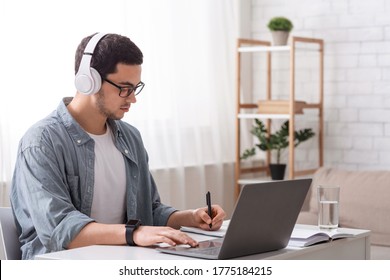 Modern gadgets for work and study online. Guy with glasses and big headphones works on laptop and makes notes in notebook in living room interior, copy space - Shutterstock ID 1775184215