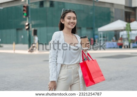 A modern gen z asian female office employee smiles while walking around the city district. Holding a cup of coffee and some shopping bags.