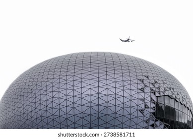 Modern geodesic dome stadium with triangular metal panels and geometric pattern, airplane flying overhead against cloudy sky in Hong Kong