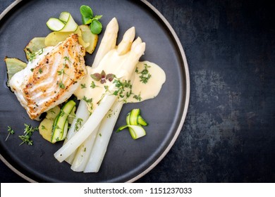 Modern German Fried Cod Fish Filet With White Asparagus In Hollandaise Sauce With Roast Potatoes And Sliced Zucchini As Top View On A Plate With Copy Space Right 