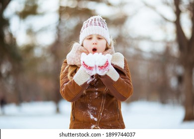 modern girl with mittens in a knitted hat and sheepskin coat blowing snow outside in the city park in winter.