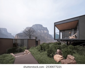 Modern glass cabin amid lush greenery, overlooking a majestic rocky mountain under a cloudy sky, with a wooden pathway leading to the entrance. - Powered by Shutterstock