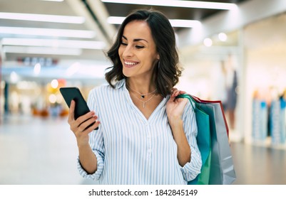 Modern Gorgeous Young Smiling Woman In Trendy Stylish Clothes With Bright Colorful Shopping Bags Is Using Her Smart Phone While Walking In The Mall