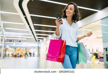 Modern Gorgeous Young Smiling Woman In Trendy Stylish Clothes With Bright Colorful Shopping Bags Is Using Her Smart Phone While Walking In The Mall