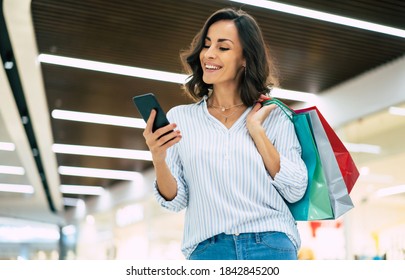 Modern Gorgeous Young Smiling Woman In Trendy Stylish Clothes With Bright Colorful Shopping Bags Is Using Her Smart Phone While Walking In The Mall