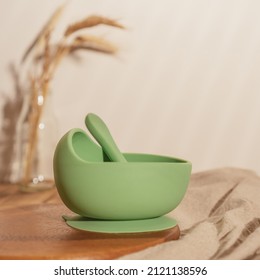 Modern green silicone bowl with suction base and spoon on wooden table near glass vase with dried flowers. Serving food, baby tableware, first feeding concept. Instagram use, square frame, soft focus. - Shutterstock ID 2121138596