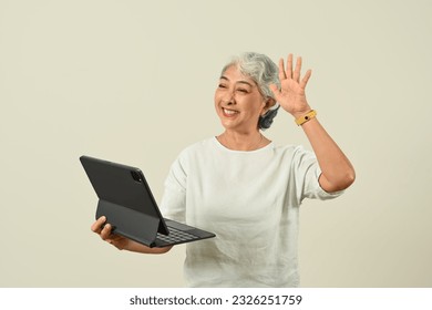 Modern grey haired woman waving hand, making video call on tablet, standing isolated on beige background - Powered by Shutterstock