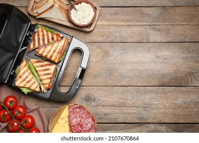 Modern grill maker with sandwiches and different products on wooden table, flat lay. Space for text