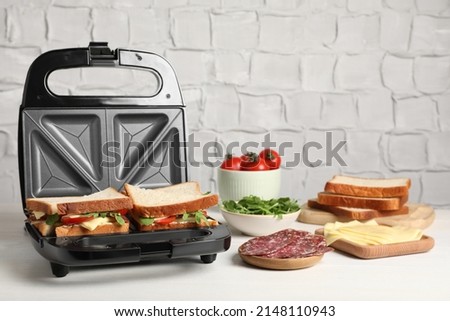 Modern grill maker with tasty sandwiches and ingredients on white wooden table
