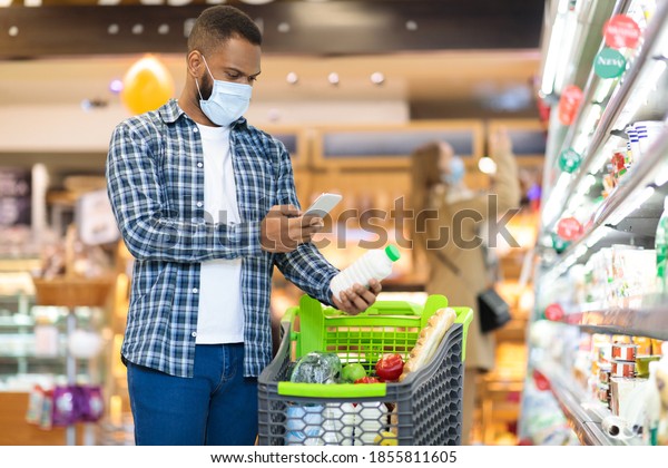 Modern Grocery Shopping. African American Man Using\
Mobile Phone Scanning Product With Smartphone App Standing With\
Trolley In Supermarket. Groceries Checklist Application. Free Space\
For Text