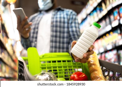 Modern Grocery Shopping. Black Man In Mask Using Smartphone Buying Food Groceries Standing Wih Trolley Cart In Supermarket Store. Customer Choosing Dairy Products In Shop. Selective Focus, Cropped