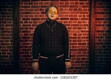 A Modern Guy With Red Hair And Short Beard Wearing A Casual Sports Suit And Stylish Glasses Stands In A Loft Style Room In Red Lighting. Youth Culture. Hip-hop Fashion. Men's Youth Style.