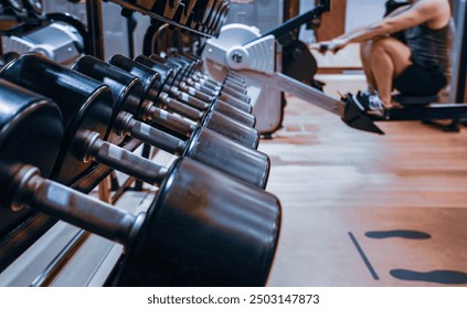 A modern gym setting with a focus on a rack of dumbbells in the foreground, with blurred gym equipment and a person working out on a rowing machine in the background. 