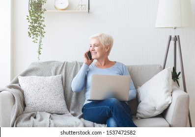 Modern Happy Old Lady Sitting On Sofa At Home, Holding Laptop And Talking By Phone, Free Space