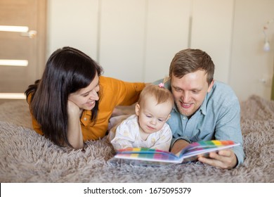 Modern Happy Young Family Spends Time Together. Mom And Dad Are Reading A Book To A Child. Literature Early Development Of The Baby. Parenting. Quarantine Self-isolation. Learning In Game Activities 