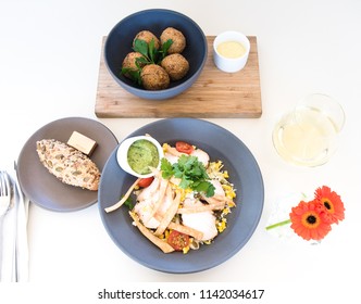 Modern Healthy Diet Tabletop Of Asian American Food And Red Gerbera Daisy Flowers. Thai Minced Meat Balls, Grilled Chicken Breasts On Mexican Salad With Avocado Dip, Bread Roll And Glass Of White Wine