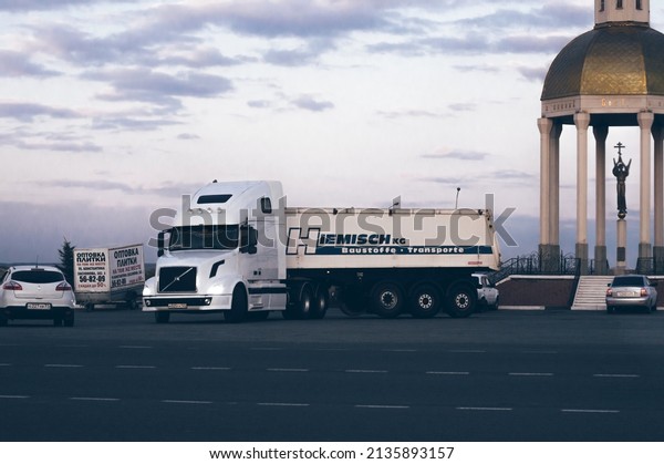 Modern heavy-duty truck for transportation with\
high cab turns onto highway with commercial cargo. Parking lot for\
cars and chapel with statue of angel at sunset. October 8, 2019,\
Belgorod, Russia