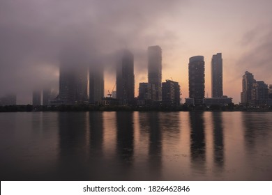 Modern High Rise Condo And Business Buildings At Waterfront Covered With Low Clouds, Fog, Haze, Smoke At Sunrise Sunset Hour Reflecting In Still Water Surface. Hot Real Estate Market Concept.