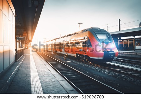 Modern high speed red commuter train at the railway station at sunset. Turning on train headlights. Railroad with vintage toning. Train at railway platform. Industrial landscape. Railway tourism