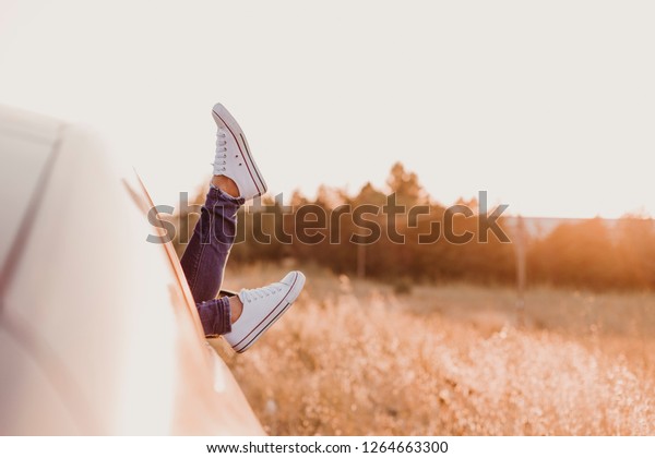Modern hipster girl resting in a car and admiring\
view. Woman driver watching sunset put feet on car door. Feet\
outside the window at\
sunset