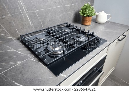 Modern hob gas stove made of tempered black glass using natural gas or propane for cooking products on light countertop in kitchen interior with oven tea pot and flowers.