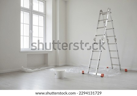 Modern home after repairs and renovations. New empty house interior with white freshly painted walls, step ladder, paint can, protective plastic on the floor, big window, and no people