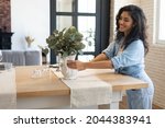 Modern home interior design, Scandinavian style concept. Beautiful young brunette woman arranging plants in vase, decorating dining room table, creating cozy atmosphere in her apartment, copy space