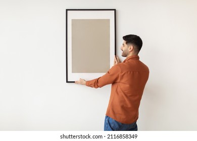Modern Home Interior And Domestic Decor. Smiling young man hanging painting, putting photo picture frame on the wall. Casual guy holding showing empty poster, design mockup, blank free copy space - Shutterstock ID 2116858349