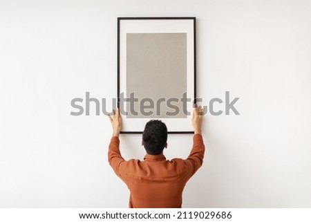 Modern Home Interior And Domestic Decor. Rear back view of man hanging painting, putting photo picture frame on white wall. Casual guy holding showing empty mock up poster, blank free copy space